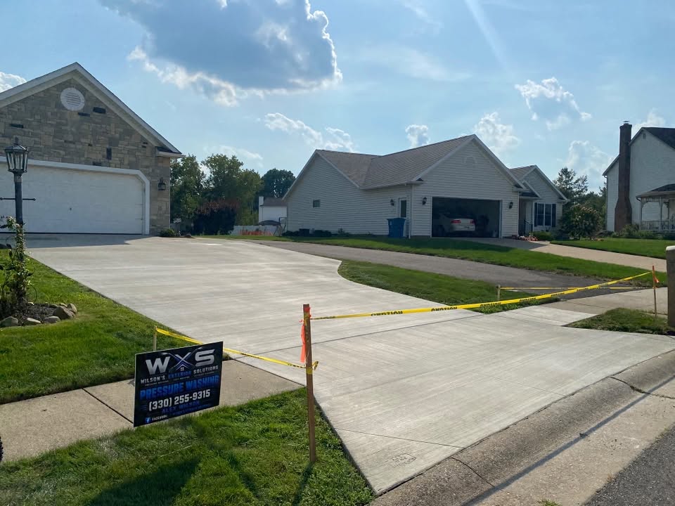 Wide concrete driveway running up to a stone-fronted house with a white garage door, freshly cleaned, a Wilson's Exterior Solutions yard sign at the edge of the lawn