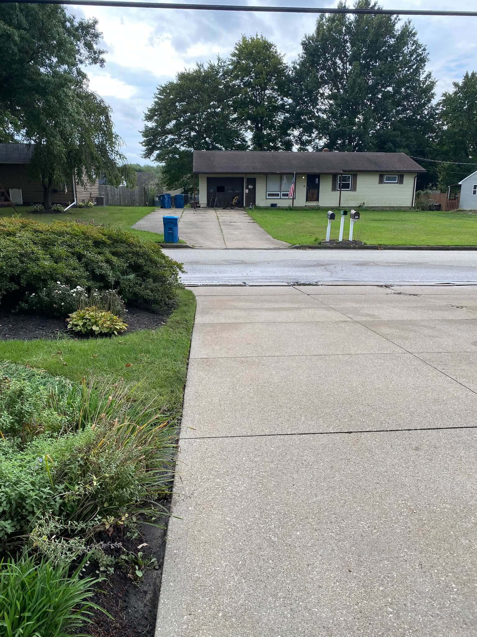 Cleaned concrete driveway running all the way down to a wet street, with a neighboring ranch house opposite