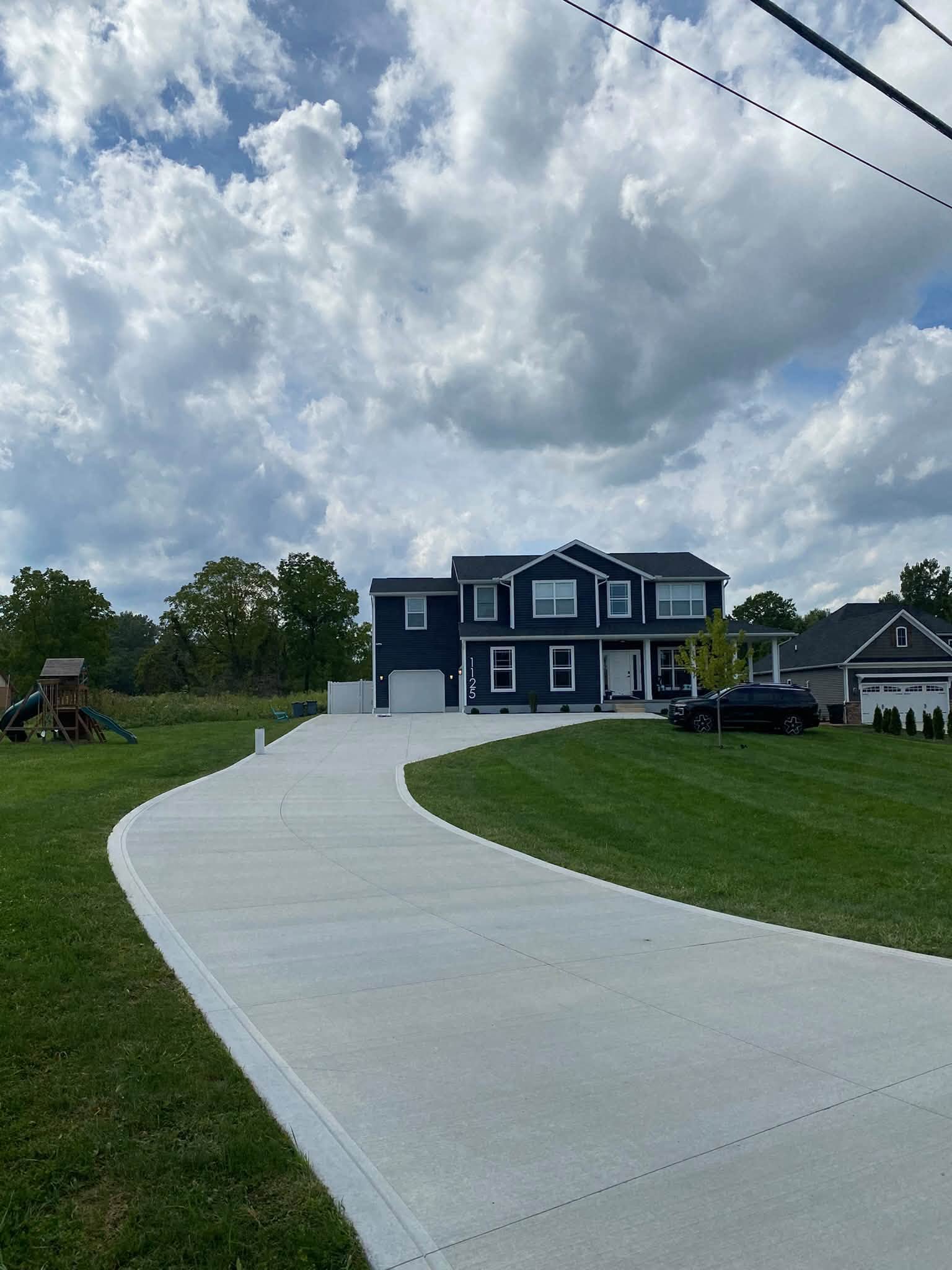 Long pale concrete driveway curving from the road up to a navy two-story home under a big cloudy sky