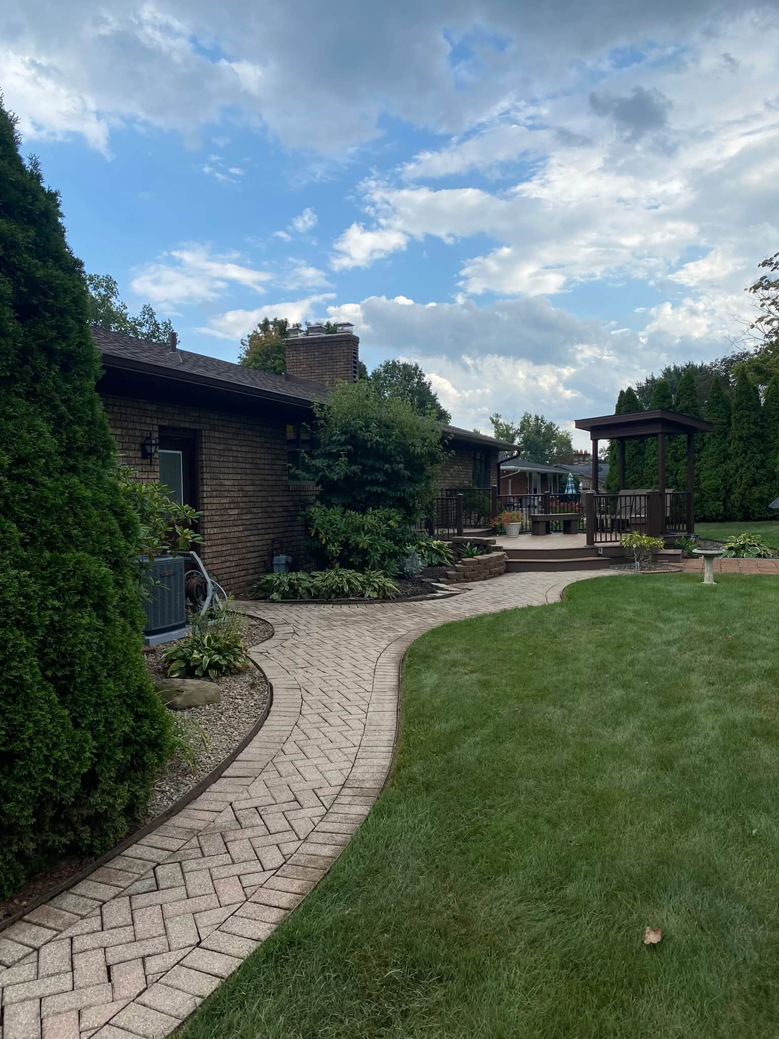 Brick paver path running alongside a brick ranch house to a raised composite deck in the evening light